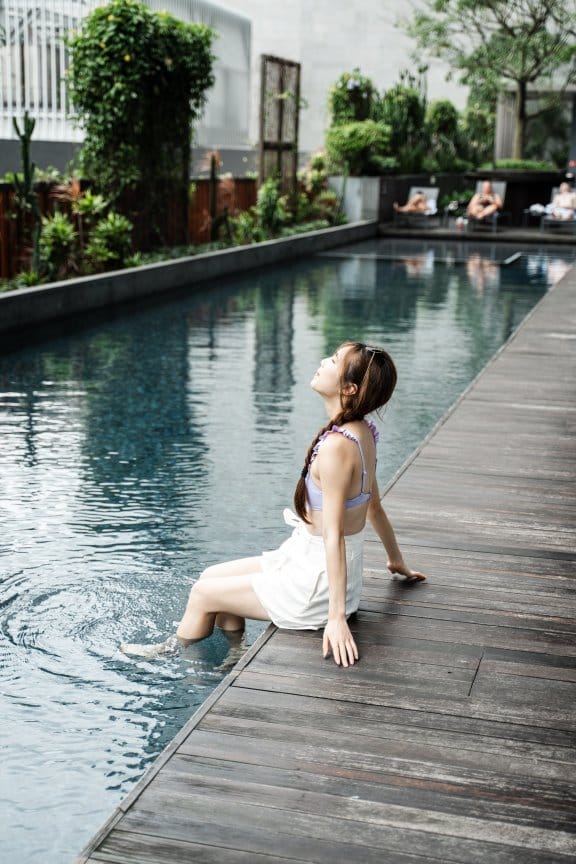 A woman dips her feet in an ourdoor hotel pool, surrounded by greenery