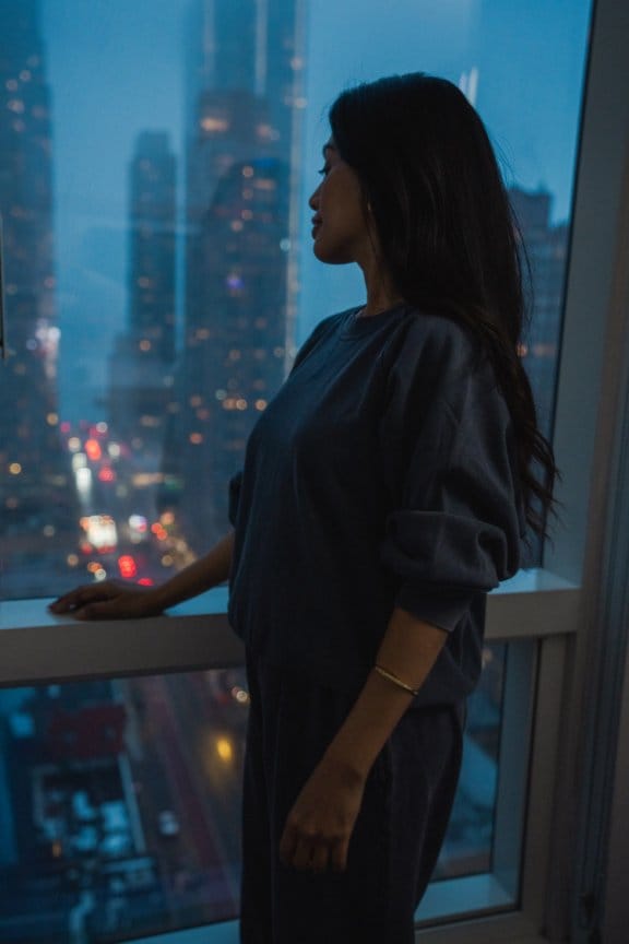 A woman stands by a large window and looks out at the New York skyline