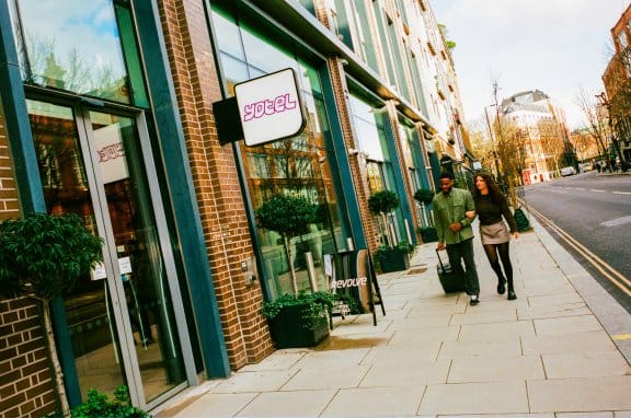 A couple walk down a sunny street approaching the front of a hotel lobby