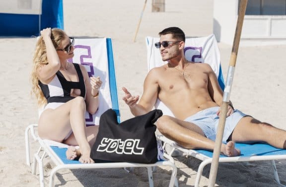 A woman and a man relax on the beach with a YOTEL tote bag