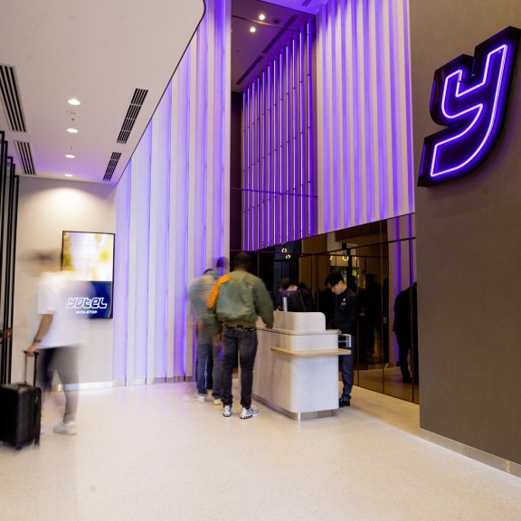Clean hotel lobby with white floors and purple panneling. The Y Yotel logo hands luminescent on the wall