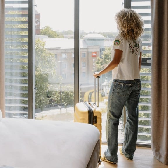 A woman with curly hair holds her suitcase and looks out her hotel room window