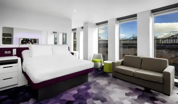 Yotel First Class bedroom with a green chair, brown sofa, green footstool/table and purple and white bed with a chest of drawers to the side of it. There are huge windows and a wide mirror 