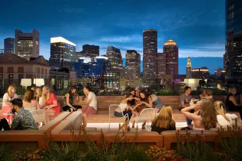 Rooftop bar at night filled with guests with the Boston skyline behind