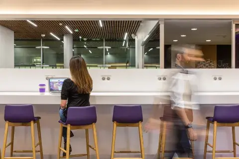 A woman in an airport hotel lounge uses her laptop as a blurry man walks by