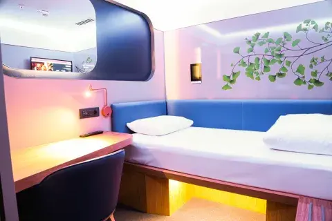 A compact hotel room with a single bed, plugs, a desk, large mirror, TV and storage space