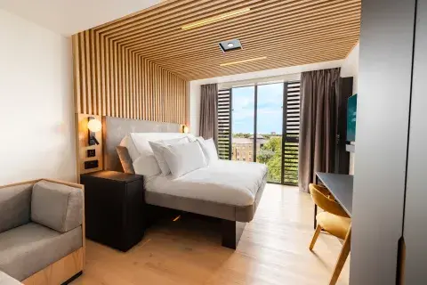 Large room with floor-to-ceiling windows, wood panelling, bedside lighting, a lounge area and a smartTV hanging from the wall.