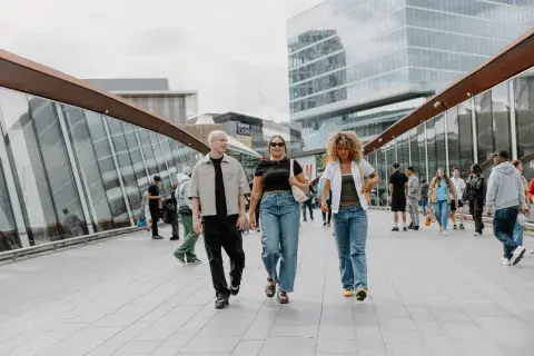 Three young adults in trendy clothes smile as they cross a modern bridge, with glistening high-rise buildings in the background