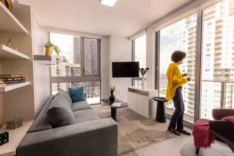 A woman in a yellow shirt looks out the window from her spacious hotel room living area, with floor-to-ceiling windows, a sofa, shelf space and a TV