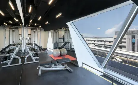 Modern hotel gym with weight machines, benches, and exercise balls beside a large angled window overlooking an airport runway with a plane taking off in the distance.