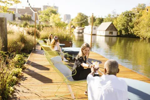 Two people clinking glasses on a sunny canal side terrace