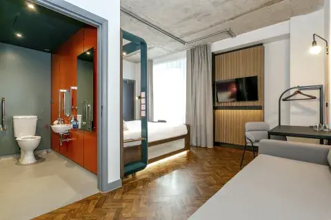 A compact hotel room with wood flooring, a large bed and red walls in the bathroom