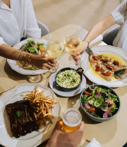Three people clinking glasses of beer and wine over a selection of fresh food