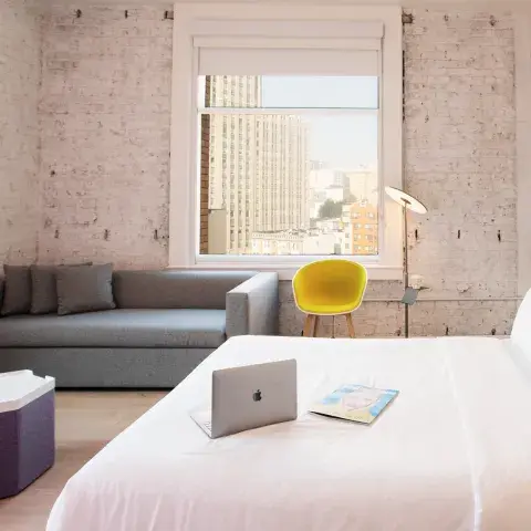A bright and airy hotel room with a sofa and large windows. A laptop and a magazine are placed on the bed.