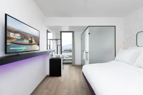 A brightly lit hotel room with white fitted sheets and a large flat-screen TV. At the end of a room, there's a large window with mountain views. 